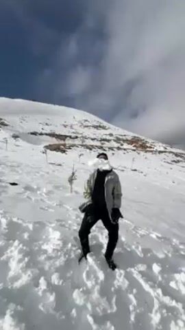 A man is standing on a snowy hillside on a sunny day. He is wearing black pants, a jacket with light-colored sections, and gloves. He throws snow into the air above him. He then bends over and picks something up from the snow. The hillside is covered with snow and there are some small trees planted along it. The sky is blue with some clouds.