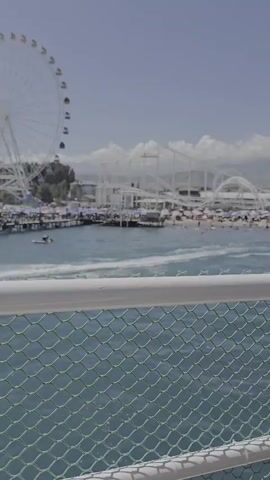 The video shows a view of a beach and amusement park across a body of water. In the foreground, a green mesh fence with white top and bottom rails is visible. The water is choppy and a jet ski is visible in the distance. The beach is populated with people and umbrellas. Behind the beach, there is an amusement park with a Ferris wheel and roller coaster. The sky is clear and blue.