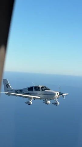 A small, white airplane with the tail number N219EL flies through a clear blue sky over a calm ocean. The plane has two visible occupants in the cockpit. The perspective is from another aircraft flying alongside. The LA flight academy logo is visible on the tail of the airplane.