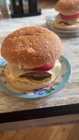 The video shows two hamburgers on plates on a table. The camera pans from one hamburger to the other. The table appears to be a kitchen or dining table. In the background, there are some kitchen appliances and a door. The hamburgers appear to be freshly made. The lighting is natural and the video is shot from a slightly high angle.