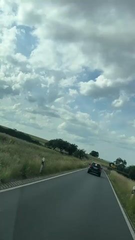 A black SUV drives along a rural road with green fields and trees on either side. The sky is partly cloudy with white clouds against a blue sky. The road has white lane markings.