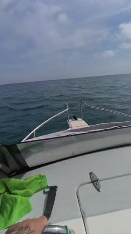 A boat moves through the water on a slightly cloudy day. The camera is positioned inside the boat, looking forward over the bow. The ocean stretches out ahead, with gentle waves visible. The sky above is a mix of blue and white clouds. A green cloth is seen on the deck of the boat in the lower left corner.