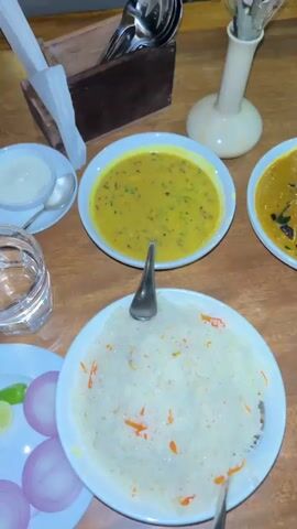 The video provides an overhead view of a wooden dining table set with a meal. Four white bowls filled with food are centrally placed: a yellow lentil soup with green seasoning, a brown curry, a plate of white basmati rice with orange strands, and an orange-red creamy curry with white swirls. Accompanying dishes include a small white bowl of white yogurt, and a small white plate holding sliced red onions and a lime wedge. A clear glass of water is also on the table. In the background, a white ceramic vase with artificial flowers, a wooden box containing various cutlery items, and a white napkin holder are visible. The camera slightly pans right across the table during the short clip.