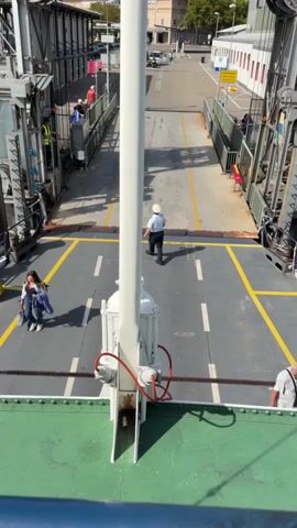 The video begins with a view from a ferry deck, looking towards the shore. People are boarding the ferry, with some walking up a ramp and others standing near the entrance. A uniformed crew member stands on the deck, seemingly directing traffic. The camera then pans to show the water and the dock area, with more people and buildings visible in the background. The scene shifts to the interior of the ferry, showing wooden benches and tables arranged on the deck, suggesting it is set up for passengers. The video concludes with a closer view of the seating area, highlighting the simple, functional design of the ferry's passenger accommodations.