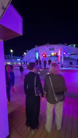 The video shows a night scene on what appears to be a cruise ship deck. Two women, viewed from behind, are holding hands and walking towards a brightly lit stage where a band is performing. The deck is illuminated by purple and pink lights, casting a colorful glow on the wooden floor. In the background, other people can be seen walking around and near the stage area. The overall atmosphere suggests a lively evening entertainment setting.