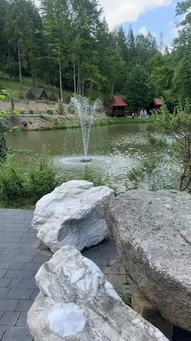A fountain sprays water in a lake surrounded by lush green trees. In the foreground, large white and gray rocks sit on a paved walkway. Red-roofed structures are visible in the background, nestled among the trees. The sky is partly cloudy.