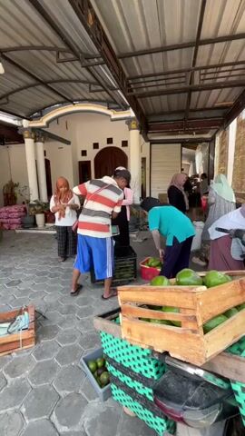 The video shows an outdoor market scene under a covered awning. Several people are engaged in transactions involving produce, primarily avocados. In the foreground, a man in a striped shirt and blue shorts holds a white sack, appearing to hand it over. To his right, a woman in a turquoise shirt and dark pants is placing avocados from a red basket into a black crate on a scale. Another woman in a pink top and hijab stands near the scale, holding a phone or tablet. Behind them, more people are present, some appearing to be customers and others working. Wooden crates filled with avocados are visible throughout the scene, as well as a motorcycle on the right side. The ground is paved with hexagonal tiles. The background features a building with columns and a large wooden door.