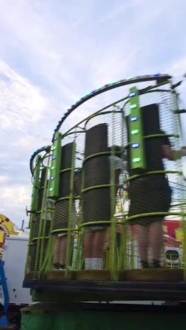 This video captures a dynamic scene at a carnival or fair. The primary subject is a green, cage-like amusement ride, possibly a spinning or swinging attraction. Within the ride, several individuals can be seen, some standing and others seemingly in motion, their figures blurred by the movement of the ride and the camera. The ride itself is adorned with a string of blue and green lights that follow its curved structure.

In the background, other carnival elements are visible. To the left, part of another ride or attraction with yellow lettering is present, along with a white trailer. Several colorful flags, red and yellow, flutter in the breeze, adding to the festive atmosphere. The time of day appears to be late afternoon or early evening, given the soft, diffused lighting and the presence of clouds in the sky. The weather seems pleasant, with a partly cloudy sky suggesting a mild day.

The overall setting is clearly an outdoor amusement park or fairground. The presence of the rides, flags, and the general ambiance points to a lively and energetic environment, likely a place for entertainment and recreation. The camera perspective is low and tilted upwards, giving a sense of the ride's scale and the sky above. While no explicit emotions are conveyed, the activity suggests excitement and enjoyment typical of a fairground experience. There is text visible on the yellow attraction to the left, but it is mostly obscured and unreadable in this footage.
