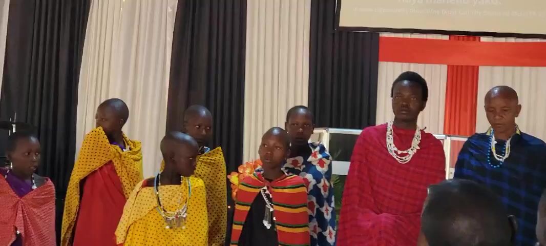 A group of Maasai people are standing in front of a black and white curtain, and a wall with a red and white checkered pattern. Several young people in the group are wearing yellow and red patterned wraps, and beaded necklaces. An older man stands to the right, wearing a blue and black plaid wrap and a beaded necklace. Another person in the group is wearing a red plaid wrap and a white beaded necklace. The heads of people in the audience can be seen in the foreground. A projector screen is visible in the upper right corner of the image.