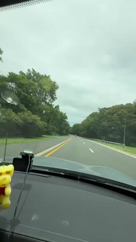 This is a first-person perspective video from inside a car traveling on a two-lane road. The road is surrounded by lush green trees on both sides, and the sky is overcast. The car's dashboard is visible in the foreground, with a dashcam mounted on the windshield. The vehicle is in motion, and the road ahead curves gently to the left. There are other cars visible in the distance. The audio includes spoken text about accidents and injuries, directing viewers to justice now dot com.