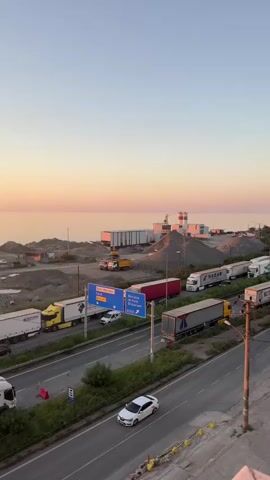 The video shows a traffic jam of trucks and cars on a highway next to the ocean at sunset. The sky is a gradient of orange and pink. There are piles of gravel and construction materials on the side of the road, as well as some industrial buildings. The trucks are lined up on the highway, indicating a significant delay. Some cars are also present in the traffic jam. The overall scene suggests a busy port or industrial area experiencing congestion.