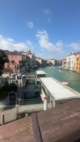 The video shows a view of Venice, Italy, from a bridge. The camera starts by looking out at a canal with colorful buildings lining the sides. Several boats are seen on the water. The camera pans to the right, showing tourists walking across the bridge. The camera is then pointed at a woman in a white and black jacket, who is standing on the bridge and waving her arms. The sky is blue with scattered clouds.
