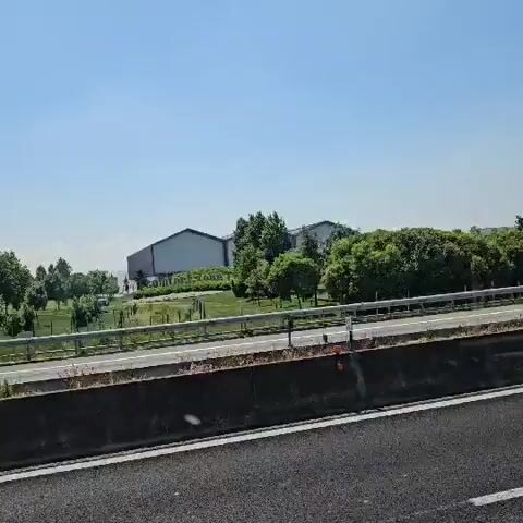 The video shows a roadside view from a vehicle traveling on a highway. The sky is clear and blue. On the other side of a barrier, there is a line of trees and bushes. Behind the greenery, there are several large, light-colored, industrial-type buildings. Cars and a truck pass on the other side of the barrier. The camera appears to be in a moving vehicle.