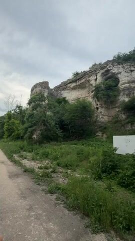 The video pans across a rocky cliff face covered in greenery. In the foreground, a dirt path runs along the bottom of the frame. A white rectangular object, possibly a sign, is visible amongst the foliage on the right side of the path. The cliff has several openings and natural formations, suggesting it may be a cave or an ancient structure. The sky is overcast.