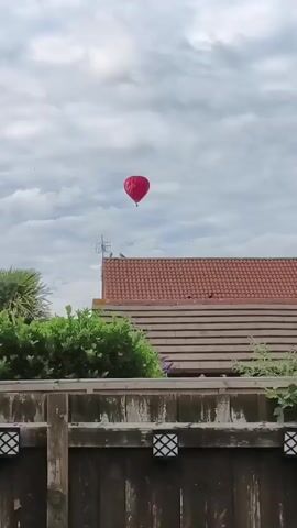 The video shows a red heart-shaped hot air balloon in a cloudy sky. Below the balloon, there is a tiled roof of a building and green foliage. In the foreground, there is a wooden fence with lights attached to it. The hot air balloon appears to be the only object in motion, slowly drifting across the sky. The sky is overcast with several clouds. The overall scene is a residential area on a cloudy day.