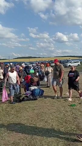 The video shows a large outdoor gathering of people carrying luggage and camping gear. The environment appears to be a grassy field under a partly cloudy sky, with a fence line and what looks like farmland in the background. Many individuals are seen walking, some with large backpacks, duffel bags, and rolled-up sleeping mats. There are several stacks of luggage visible. The overall impression is of a group preparing to camp or depart from a location like a festival or campsite. The people are casually dressed, suggesting warm weather. There are no obvious signs of distress or conflict.