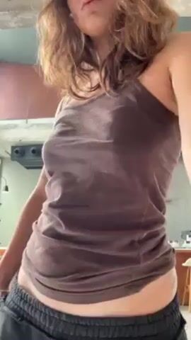 The video shows a young woman indoors, with dark hair, wearing a brown tank top and loose, dark-colored sweatpants. She is standing in front of a table or counter with various small bottles and objects displayed on it. Behind her are a couple of men, one wearing an orange and white striped shirt and the other wearing a white shirt. The woman runs her hand through her hair and then turns around to look at the camera, briefly revealing a tattoo on her lower back. She has a white bag hanging over her shoulder. The background shows a kitchen area with a vent hood and simple decor. The lighting appears natural.