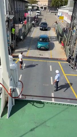 A video taken from a higher vantage point shows a ferry ramp with several cars and people. In the foreground, the green deck of the ferry is visible. Two men stand on the ferry's deck. One man, wearing a white t-shirt and dark shorts, stands facing away from the camera. The other man, wearing a white uniform with a hat, stands facing the camera. A turquoise car is positioned between them, facing away from the camera. Further up the ramp, a black car is approaching the turquoise car. Beyond the black car, there are more people and vehicles on the ramp and in the background, suggesting a port or terminal environment. The ramp is bordered by metal railings and structures. Sunlight illuminates the scene, casting shadows on the ground.