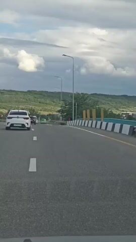 The video shows a point-of-view from inside a vehicle traveling on a highway. The highway has multiple lanes and is bordered by a barrier with black and white diagonal stripes and some yellow posts. Streetlights line the side of the road. In the distance, there are green hills and a cloudy sky. Several other vehicles are visible on the highway, including cars and a truck. One car in the foreground is dark-colored, and a white car is ahead of it. Another car further ahead appears to be white. The video is taken during daylight hours.