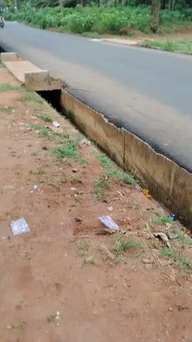 The video captures a daytime scene in Akwa Etiti, Nigeria, showing a paved road with a person riding a motorcycle in the distance. The road appears to be a two-lane asphalt path, flanked by a concrete drainage ditch on the right side. The ground adjacent to the ditch is reddish-brown earth, littered with several pieces of plastic waste and wrappers. Beyond the road, dense vegetation, including numerous palm trees and other green foliage, lines both sides, suggesting a rural or semi-rural setting. The sky is overcast or partly cloudy, providing diffused light without strong shadows. The person on the motorcycle is moving away from the camera, gradually becoming smaller as they proceed down the road.
