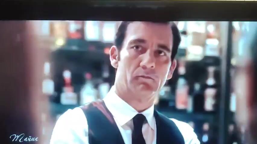 A man in a white shirt and black vest, with dark hair, looks to his left. He has a serious expression. In the background, there are bottles on shelves. Then the scene changes to a woman in a red dress with long, wavy brown hair. She is looking down. Behind her, several figures are dancing in dimly lit, beige-colored room. To her right, a photographer with a camera is visible. In the foreground, men are seated, watching the scene. The woman turns her head slightly to her right.