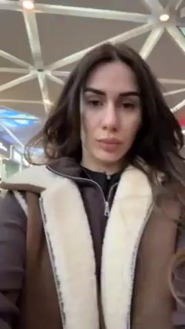 A woman with long brown hair is centered in the frame. She is wearing a brown jacket with a white fuzzy lining and brown suede exterior. She makes a kissing face at the camera. She appears to be indoors in a large open space, possibly a warehouse or market, with boxes and a metal beam structure visible in the background. The lighting is bright and somewhat industrial. The camera appears to be handheld, at eye level, and stable.