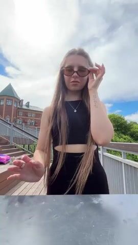 A young woman with long, dark blonde hair stands on a wooden walkway, looking towards the camera. She wears a black, sleeveless crop top and a black, form-fitting skirt. She has on sunglasses and a silver necklace. Her left hand is raised, adjusting her sunglasses. A tattoo is visible on her left forearm.

The setting appears to be outdoors, possibly a park or public area, with a railing to her right and wooden stairs to her left. In the background, a large, multi-story building with a green, conical roof is visible, along with a white bell tower. Lush green trees are also present. The sky is partly cloudy, with the sun shining brightly through a gap in the clouds, suggesting it is daytime, likely mid-morning to early afternoon. The overall mood is casual and relaxed.

There is no visible text in the media file.