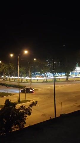 This is a night-time video clip of an intersection in Kampung Makam, Malaysia. The scene is illuminated by streetlights, which cast a warm glow on the asphalt road and surrounding trees. A dark-colored car is visible making a turn at the intersection, its red taillights a bright contrast against the darkness. In the background, several other vehicles with their headlights and taillights create streaks of light, suggesting a moderate amount of traffic. The overall atmosphere is calm and quiet, with the darkness of night dominating the scene. The trees lining the road are lush and green, adding a natural element to the urban setting. There is no visible text in the video.
