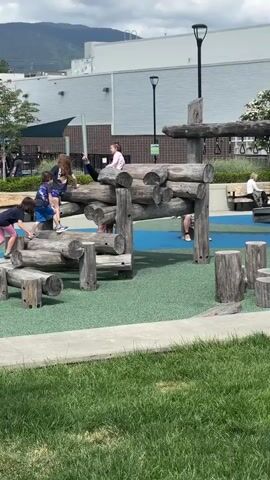 The video shows four children playing on a playground structure built of wooden logs. The structure is set on a playground surface of blue and green rubberized material. The children are climbing and crawling on the logs. There are two girls and two boys, they are all wearing casual clothing. In the background, there is a building with a corrugated metal facade and a black awning. Also in the background, there are trees, a black lamppost, and a mountain. A man wearing a white baseball cap, black collared shirt with a green logo and grey shorts walks from behind the play structure. He walks left to right on the video. The grass is green and there is a concrete walkway adjacent to the playground. It is a bright, overcast day.