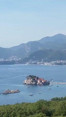 The video shows a wide aerial view of a coastal town on a sunny day. The foreground is dominated by lush green trees. In the middle ground, a small, rocky island with buildings is connected to the mainland by a narrow causeway. Several boats are visible in the blue water surrounding the island. Beyond the island, the coastline stretches out, lined with buildings and a beach. In the background, a range of hazy mountains rises against a clear blue sky. The overall impression is one of a picturesque Mediterranean landscape.