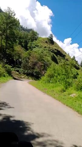 The video shows a paved road winding through a lush green hillside. The road curves to the left and appears to be going uphill. On the right side of the road, there is a steep, grassy slope covered with various trees and foliage. A utility pole with wires is visible on the right side. The sky is bright blue with fluffy white clouds. On the left side of the road, the slope is also covered with trees and vegetation. In the distance, the road continues to curve upwards along the hillside. The overall impression is of a scenic, rural landscape on a sunny day.