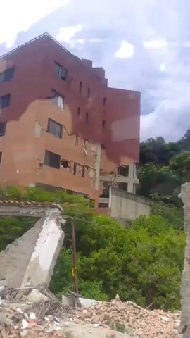 The video shows a brick building that appears to be damaged, with some sections of the facade crumbling and windows broken. Debris, including broken concrete and rebar, is visible in the foreground. Lush green vegetation surrounds the building, contrasting with the evident structural damage. The sky is overcast, suggesting a cloudy day. The camera is moving, providing a panning view of the building and its surroundings. The overall impression is one of a structure that has undergone some form of destruction or significant decay.