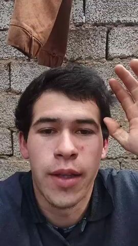 A medium shot captures a young man with dark hair and a gray collared shirt against a cinderblock wall. A brown jacket hangs in the background. The man waves, then clenches his fist. He speaks to the camera.