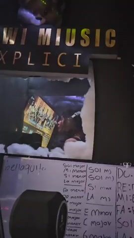 The video shows a person, likely a young woman, in a home music studio located in Alto Hospicio, Chile. She is wearing headphones and singing into a microphone, focused on recording vocals. A computer monitor in front of her displays what appears to be a video game or a graphic-intensive application featuring multiple characters.

Above the monitor, golden letters spell "I MIUSIC EXPLICIT," with a decorative, light-colored object placed on top. White fluffy material, resembling artificial clouds, surrounds the monitor, enhancing the visual aesthetic of the space. The lighting in the room changes, showing blue and green tones, indicative of dynamic LED lighting commonly used in creative environments.

In the foreground, a whiteboard displays handwritten musical notes. These include chord names and scales such as "bellaquito," "Sol mayor," "Si menor," "La mayor," "Fa menor," "DO mayor," "RE major," "MI major," and "C major," along with solfège symbols like "DO," "RE," "MI," "FA," and "SO." A studio monitor speaker is also visible, completing the recording setup. The scene depicts a dedicated and actively used music production space.