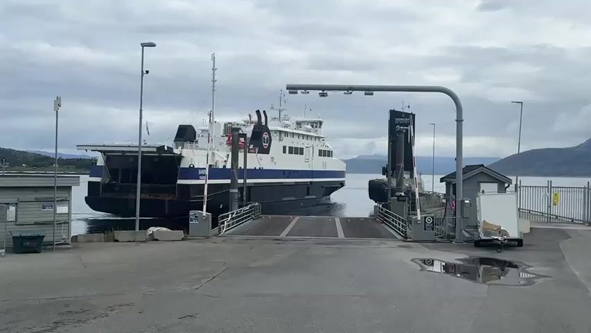 The video shows a ferry docked at a terminal. The ferry is a large vessel with a dark hull and a white superstructure. It has a ramp at the stern, which is extended to meet the dock. The dock is paved and has a few buildings and some equipment. There are mountains in the background. The sky is overcast. The ferry appears to be preparing to depart or has just arrived. The ramp is lowered to the water.