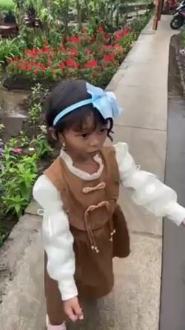 A young child with a light blue bow headband is walking down a path in a garden. The garden is full of colorful flowers. The child is wearing a brown dress with white puffy sleeves. The path is made of concrete slabs. The child glances to the side, then to the viewer. The camera follows the child as she walks up a set of stone steps. A family is walking toward a building with a gravel path in front of it. The building has a white exterior with a bamboo frame.
