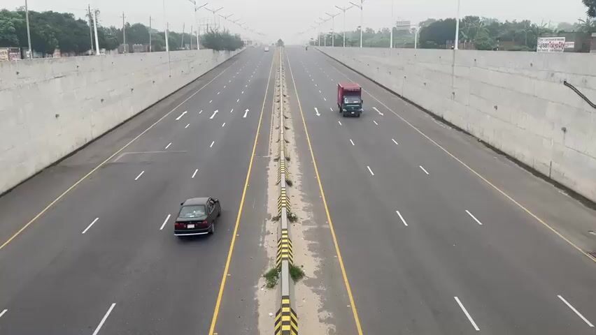 The video depicts a wide, multi-lane highway with concrete barriers on either side and a central barrier marked with black and yellow stripes. The highway surface is asphalt, and lane markings include solid yellow lines and white dashed lines. Traffic is relatively light, consisting of a few cars and trucks. Two blue trucks and a green truck travel together in the left lanes of the near side of the highway. Further down the highway, a dark car moves alone on the left lanes of the near side. A red container truck is in the right lanes of the near side of the highway. Streetlights are visible along the highway, and vegetation and buildings can be seen in the distance. The sky is overcast, creating a slightly hazy atmosphere. The camera angle is from an elevated position, likely a bridge or overpass.