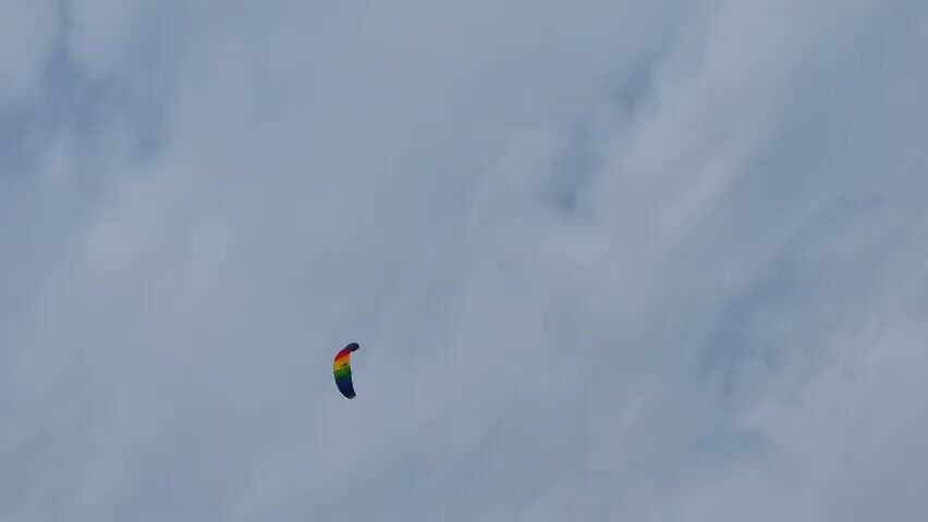 A colorful kite flies against a cloudy sky. The kite is small and appears to be a power kite with a rainbow color scheme. The sky is overcast with soft, diffused light. The bottom right corner of the frame shows the edge of a mountain. The kite is in the upper left quadrant of the frame and is moving across the sky.