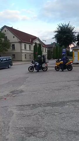Two motorcycles are visible on a paved road. The motorcycle on the left is white and silver with a rider wearing a dark helmet and jacket. The motorcycle on the right is yellow with a rider wearing a dark helmet and blue jacket. The yellow motorcycle is turning right and moving away from the camera. The white and silver motorcycle is stationary. In the background, there is a building with a red roof, trees, and a bus stop. There are also several pedestrians standing near the bus stop. The sky is cloudy.
