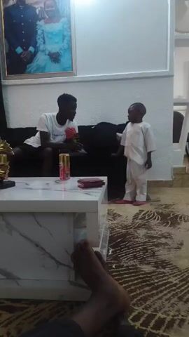 The video shows two people, one a young man and the other a young child, in what appears to be a living room. The young man is sitting on a black couch, wearing a white t-shirt with the words "HelloLabs" on it, and black shorts. He's holding a red and green toy in his hands and smiling. A can of Amstel beer and a small maroon wallet are on the white marble-patterned coffee table in front of him. The young child, wearing a white outfit, stands in front of the coffee table and looks at the young man, appearing to be engaged in a conversation. The background includes a patterned brown and beige rug, a picture hanging on the wall behind the couch, and a view of someone's feet at the bottom of the frame, indicating they may be filming the scene.