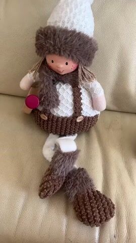A close-up shot shows a handmade doll sitting on a tan leather couch. The doll is dressed in winter attire, including a white knitted hat with a brown fur trim, a brown fur scarf, a white knitted sweater with a diamond pattern, and brown overalls with wooden buttons. Its arms and legs are visible, and it has brown yarn hair and a rosy-cheeked face with freckles. In its left hand, the doll holds a small wooden object with a pink circular top. The doll's legs are crossed, and it wears brown knitted leg warmers with fur trim. The lighting is bright, highlighting the doll's features and the texture of its clothing.