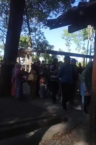 A group of people are standing in a line outdoors. The background suggests a rural or tropical environment with trees and some rudimentary structures. The people appear to be waiting for something or someone. Some are dressed in traditional clothing, while others wear more casual attire. The lighting indicates it is daytime, possibly morning or afternoon. The scene is somewhat shadowed due to overhead trees and a nearby structure.