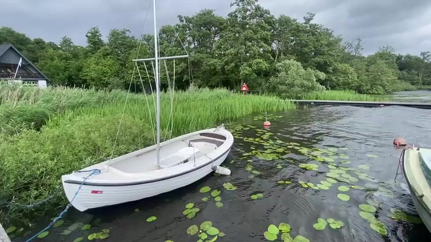 The video shows a white sailboat docked on a body of water surrounded by lush green vegetation and trees under a cloudy sky. A building with a dark roof and white walls is visible on the left side of the frame, with a flag on a pole in front of it. The sailboat is tied to a dock with a blue rope. Lily pads dot the surface of the water around the boat. Another boat, partially visible, is docked to the right of the sailboat. The overall scene suggests a peaceful, natural setting by the water.