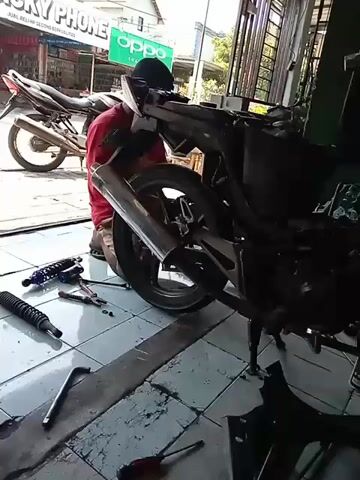 The video shows a person kneeling on the ground in front of a disassembled motorcycle. The motorcycle is missing its body panels and appears to be undergoing repairs or maintenance. Various tools, including wrenches and a screwdriver, are scattered on the tiled floor around the motorcycle. A power drill is also visible. In the background, another motorcycle is parked, and a sign for "OPPO" is displayed on a building. The scene appears to be an outdoor workshop or a roadside repair area.