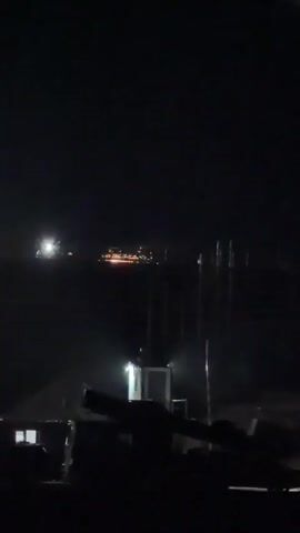 The video shows a night scene with a dark, rural environment. In the background, there is a distant line of illuminated buildings, possibly a town or city, with some lights also visible on the water. In the middle ground, there are several tall, thin vertical structures that are illuminated by an unseen light source, casting shadows and reflections on the water. Closer to the foreground, there is a small, brightly lit structure, possibly a guardhouse or control booth, with a person or figure visible inside. In the immediate foreground, silhouetted objects are visible, which appear to be large pieces of machinery or equipment, possibly military in nature, with a dark, indistinct background. The overall atmosphere is dark and somewhat mysterious, with the lighting creating a stark contrast between light and shadow.