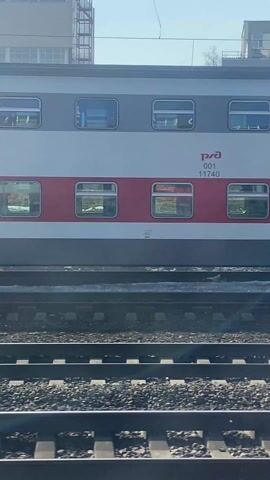 The video shows a passing train car on a railway track. The train car is white and red, with a gray stripe on the bottom and gray windows. There are numbers written on the car. The railway track is made of metal and gravel. In the background, there are buildings visible under a blue sky.