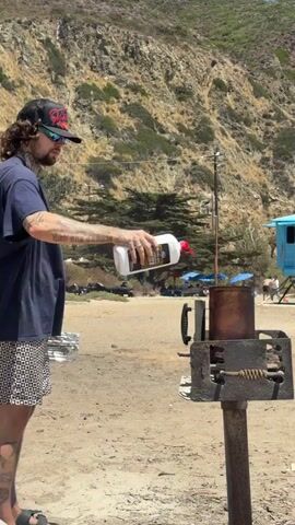 A person wearing a dark blue t-shirt, patterned shorts, a baseball cap, and sunglasses is standing on a sandy beach. They are holding a white bottle with a red cap and pouring liquid into a metal container that is on fire. The container is part of a grill, which is mounted on a metal pole. Behind the person, there is a hillside covered with dry brush and some trees. In the background, there are a few people, some umbrellas, and a small blue structure. The sky is clear and the sun is shining. The video captures the action of the person adding liquid to the fire, causing it to flare up.