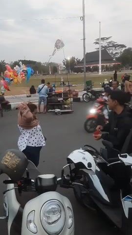 A crowded outdoor scene, possibly a park or plaza, with many people and motorcycles. In the foreground, two scooters are parked side-by-side, one white and one black. A man in a black jacket is sitting on the black scooter, holding a small red object, possibly a drink or food item. To his left, a woman in a white polka dot shirt and a scarf stands with her hands behind her head. Further back, a street vendor is visible with a cart and balloons. Many other people are scattered throughout the scene, some walking, some sitting, and others on motorcycles. A few kites are flying in the distance. A large building with a dark roof and decorative lights is in the background, with more parked motorcycles in front of it. The sky is overcast.