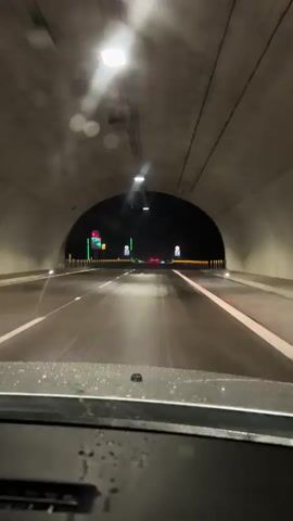 A car is driving on a highway at night. The road is wet from rain. There are lane markings and guardrails visible. Lights from the highway and signs illuminate the scene. The driver is inside the car, looking out the windshield. The video captures the view from the driver's perspective.