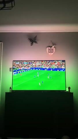 A television screen displays a soccer match. The screen is framed by green LED lighting on the sides and pink LED lighting at the top. Above the TV, a wall decoration features two butterflies and a heart-shaped sign that reads "STAY HOME LOVE". The TV is placed on a black cabinet. The lower part of the image shows the legs of a person or people.